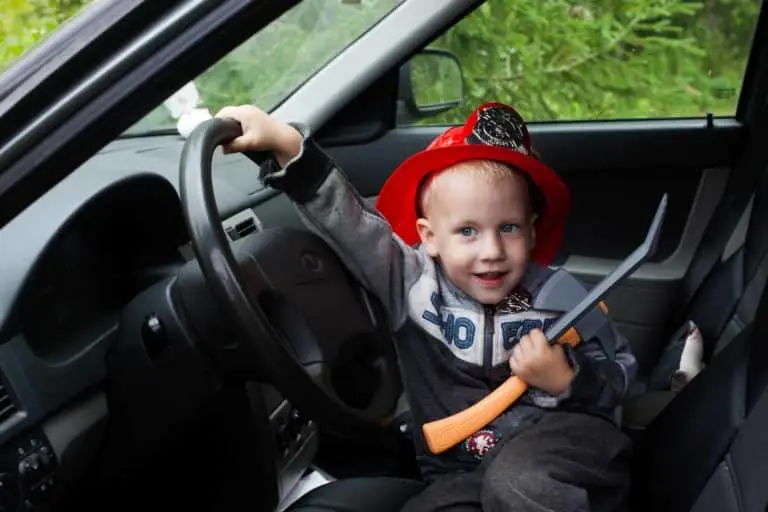 A young child with a firefighter's helmet and a toy axe playfully takes the driver's seat of a car, pretending to be a firefighter on the go.