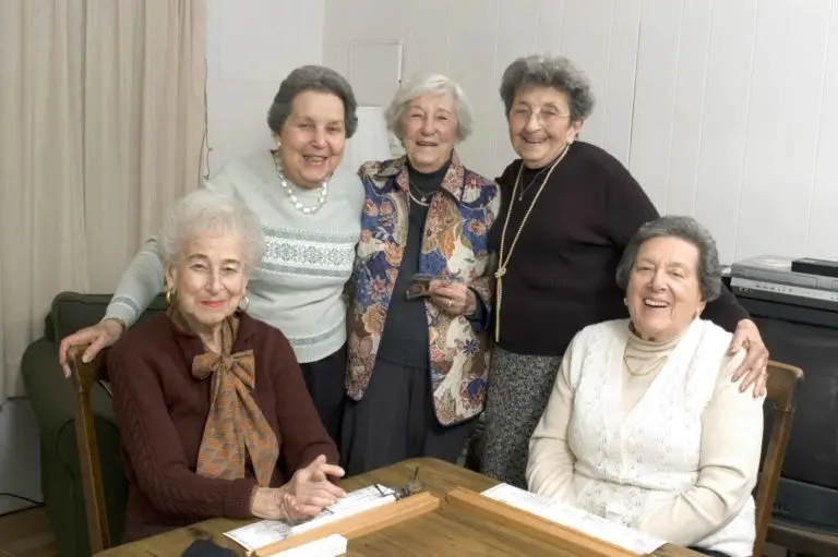 Five smiling elderly women enjoying each other's company, gathered around a table in a cozy room.