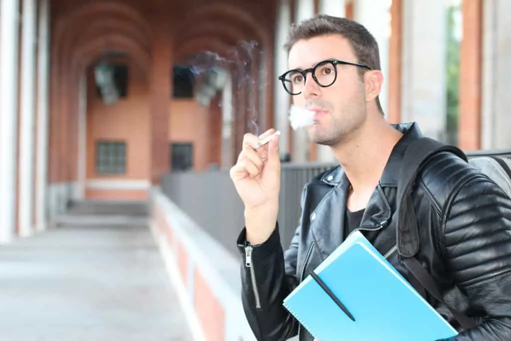 A young man in a leather jacket and glasses holding a blue notebook takes a break to smoke a cigarette, with a blurred background featuring what appears to be a covered walkway with arches.