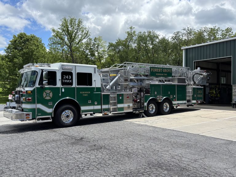 A green and white fire department ladder truck labeled “Forest Grove 243 Truck” is parked outside a fire station on a sunny day, with trees and clouds visible in the background.