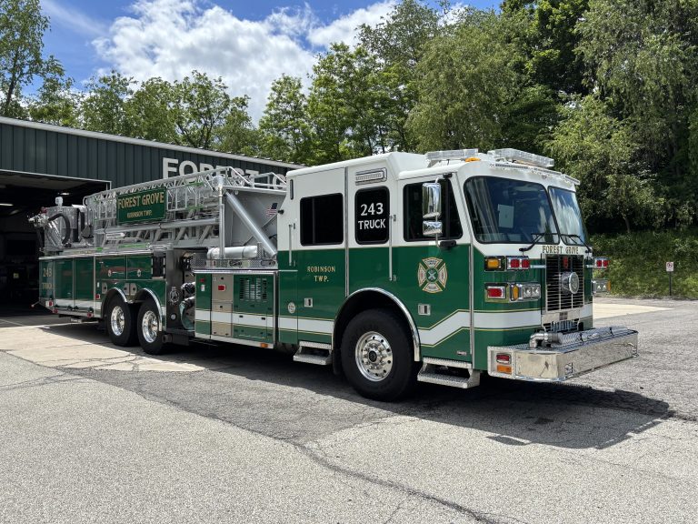 A green and white fire department ladder truck labeled “Forest Grove 243 Truck” is parked outside a fire station garage, surrounded by trees and greenery.