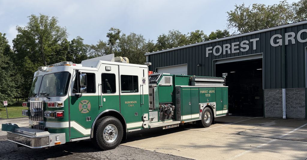A green and white fire department ladder truck labeled “Forest Grove” is parked outside a metal fire station garage, with trees and a clear sky in the background.