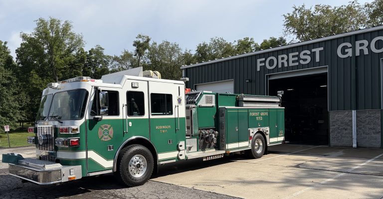 A green and white fire department ladder truck labeled “Forest Grove” is parked outside a metal fire station garage, with trees and a clear sky in the background.