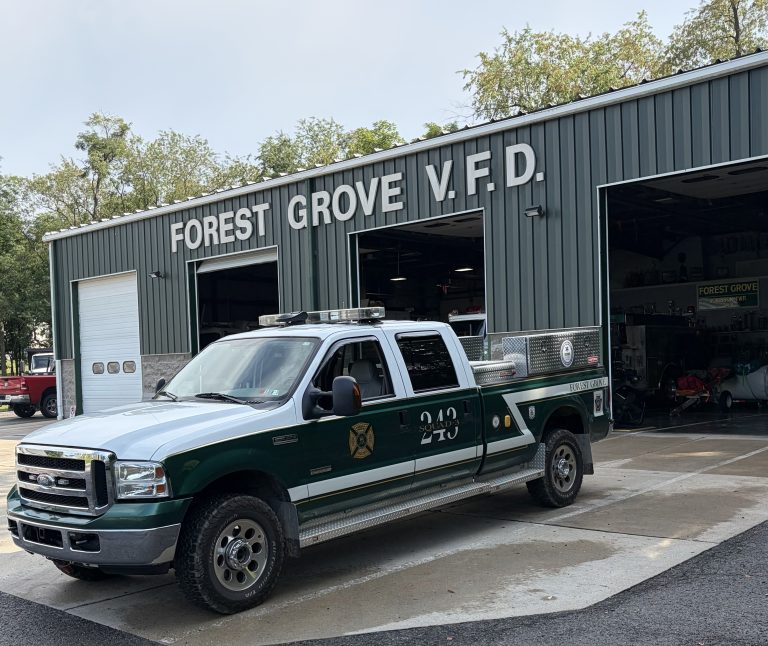A green and white fire truck labeled “243,” which serves as the fire department ladder truck, is parked outside the Forest Grove Volunteer Fire Department station, with garage doors open and equipment visible inside.