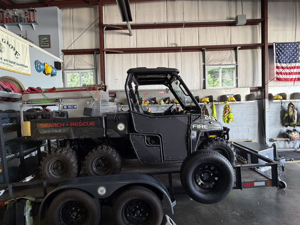 A six-wheeled search and rescue vehicle is parked on a trailer inside a fire station, with firefighting gear, helmets, and an American flag hanging on the wall in the background. Nearby, a fire department ladder truck stands ready for emergency response.