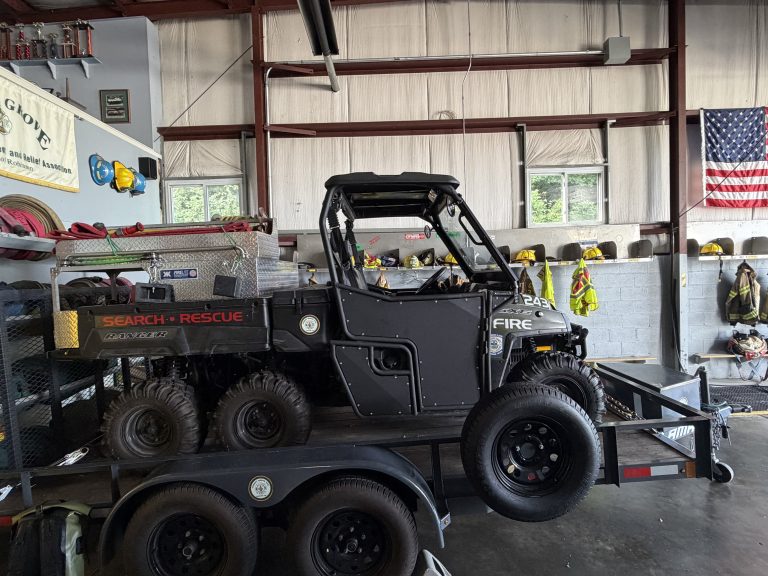 A six-wheeled search and rescue vehicle is parked on a trailer inside a fire station, with firefighting gear, helmets, and an American flag hanging on the wall in the background. Nearby, a fire department ladder truck stands ready for emergency response.