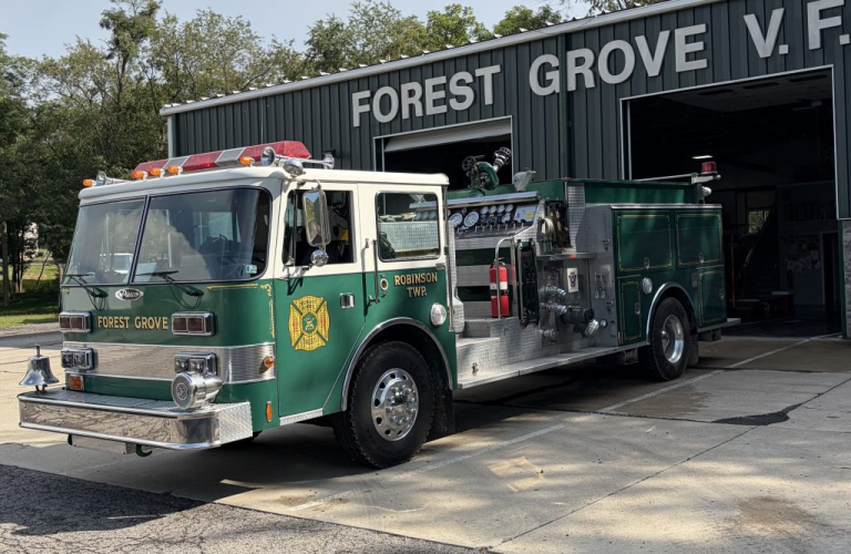A green fire engine apparatus labeled “Forest Grove” is parked outside a green metal fire station with large open doors. The building sign reads “FOREST GROVE V.F.” and trees are visible in the background.