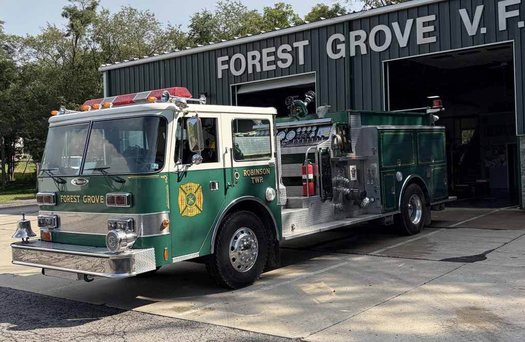 A green fire engine apparatus labeled “Forest Grove” is parked outside a green metal fire station with large open doors. The building sign reads “FOREST GROVE V.F.” and trees are visible in the background.