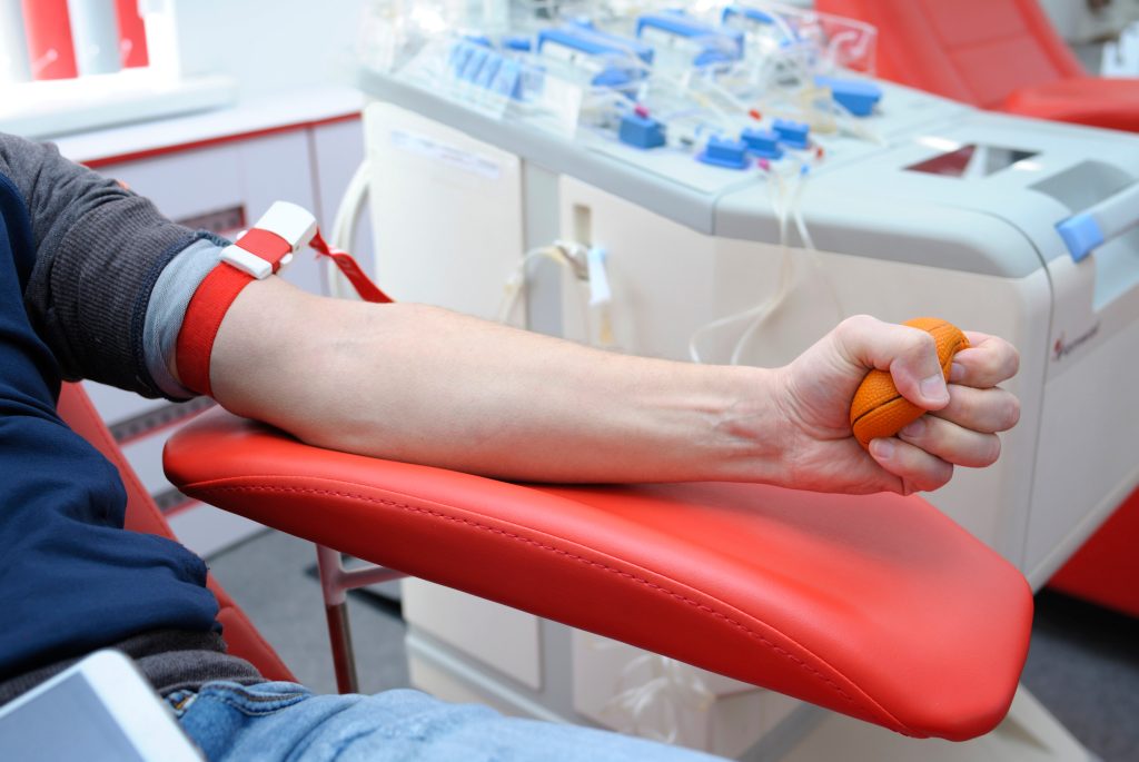 A person donates blood, squeezing an orange ball while seated in a red chair, with medical equipment and tubes attached to their arm.
