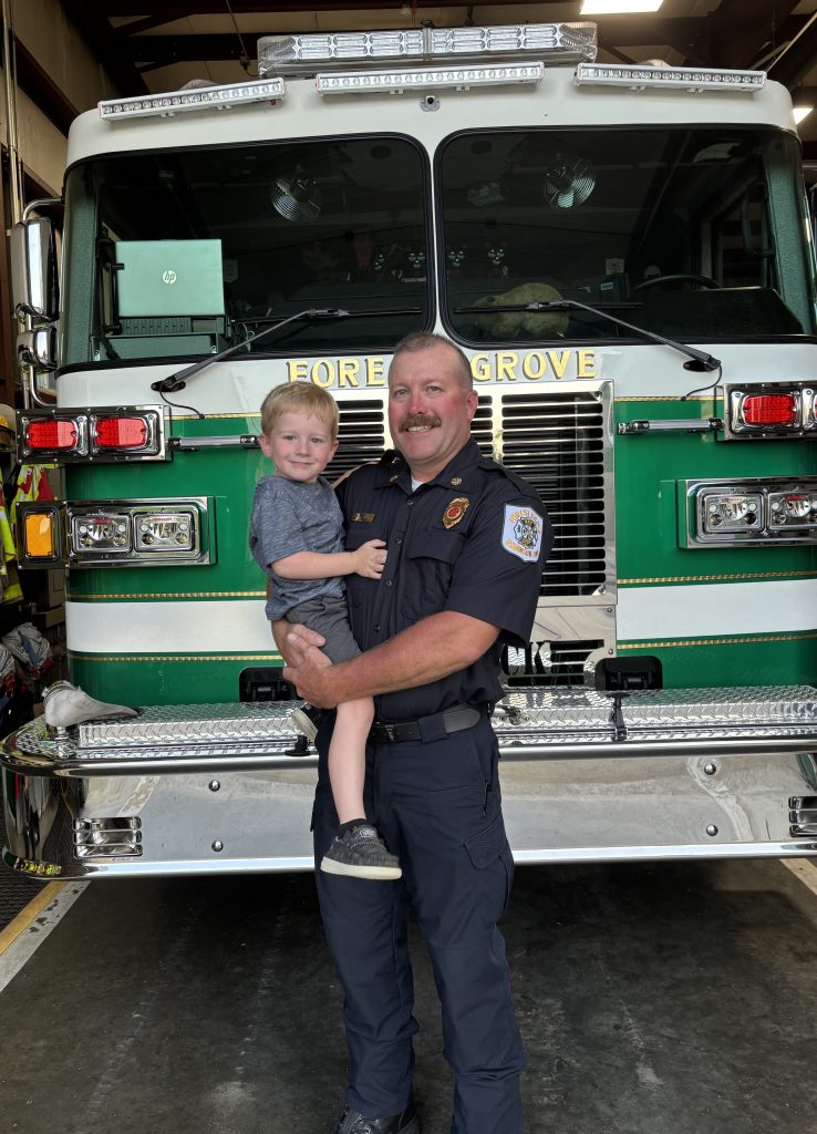 A firefighter in uniform smiles while holding a young child in front of a green and white fire truck inside a fire station.