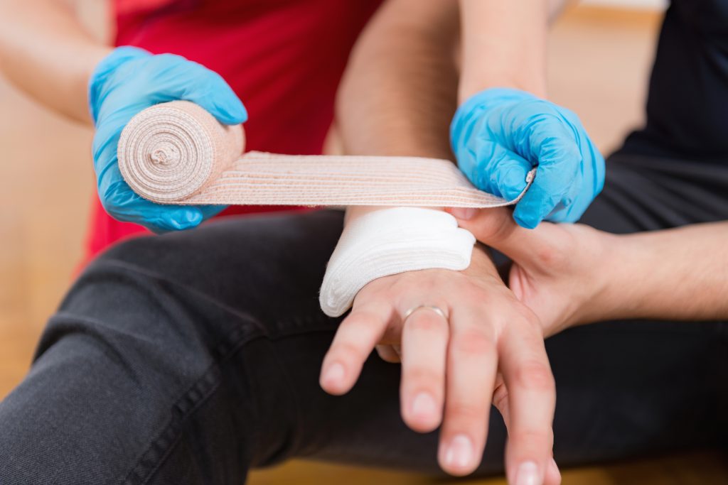 Person in blue gloves wraps gauze bandage around another person’s wrist, which already has a white pad applied, suggesting first aid care for an injury.