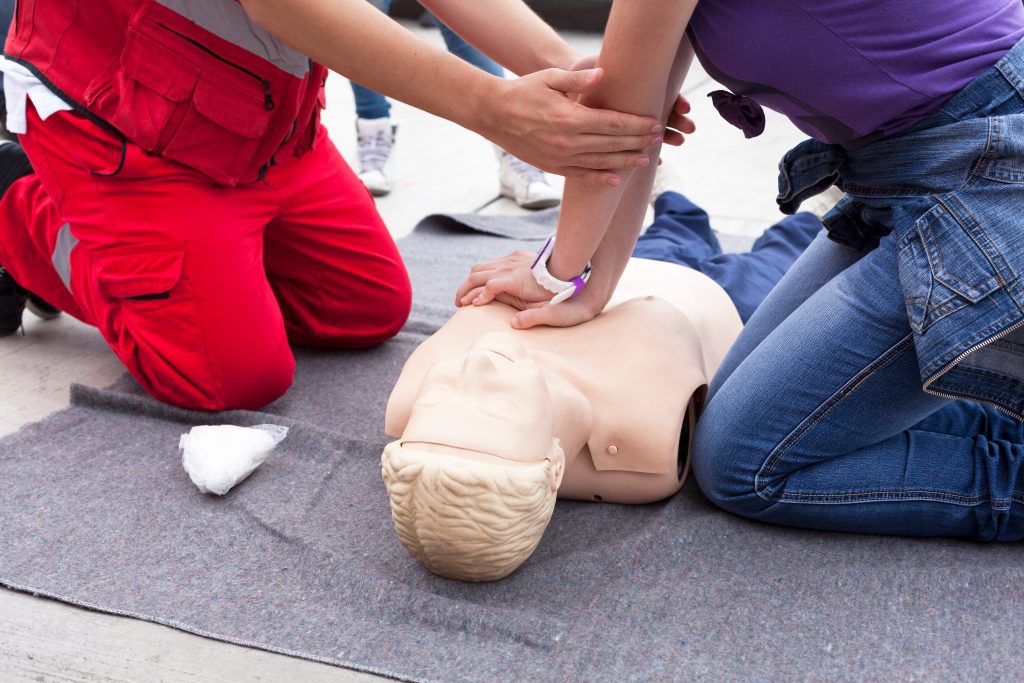 Two people kneel on a mat practicing CPR chest compressions on a training mannequin, demonstrating a first aid procedure.