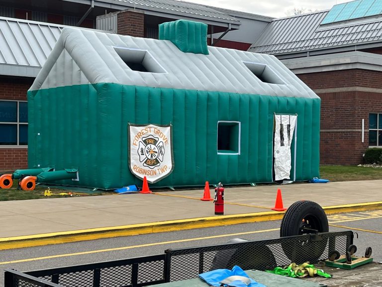 A large green-and-gray inflatable house sits outside a building. Orange cones, a fire extinguisher, and a sign reading “Forest Grove Fire” are nearby. Toys and tires are visible in the foreground.