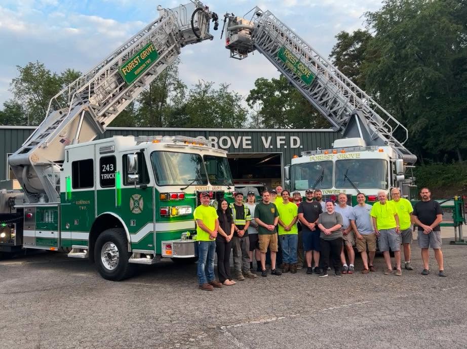 A group of firefighters and community members stand in front of two green and white fire trucks, with raised ladders, outside a building labeled “Forest Grove V.F.D.”—a proud testament to Forest Grove volunteer fire department history. Trees are visible in the background.