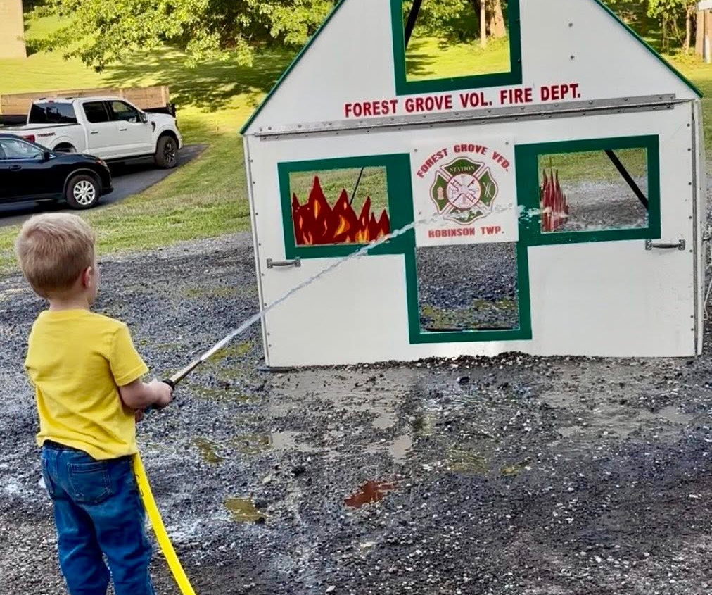 A young boy uses a yellow fire hose to spray water at a model house with painted flames, simulating putting out a fire during a fire department open house. The house displays “Forest Grove Vol. Fire Dept.” and a fire department emblem.