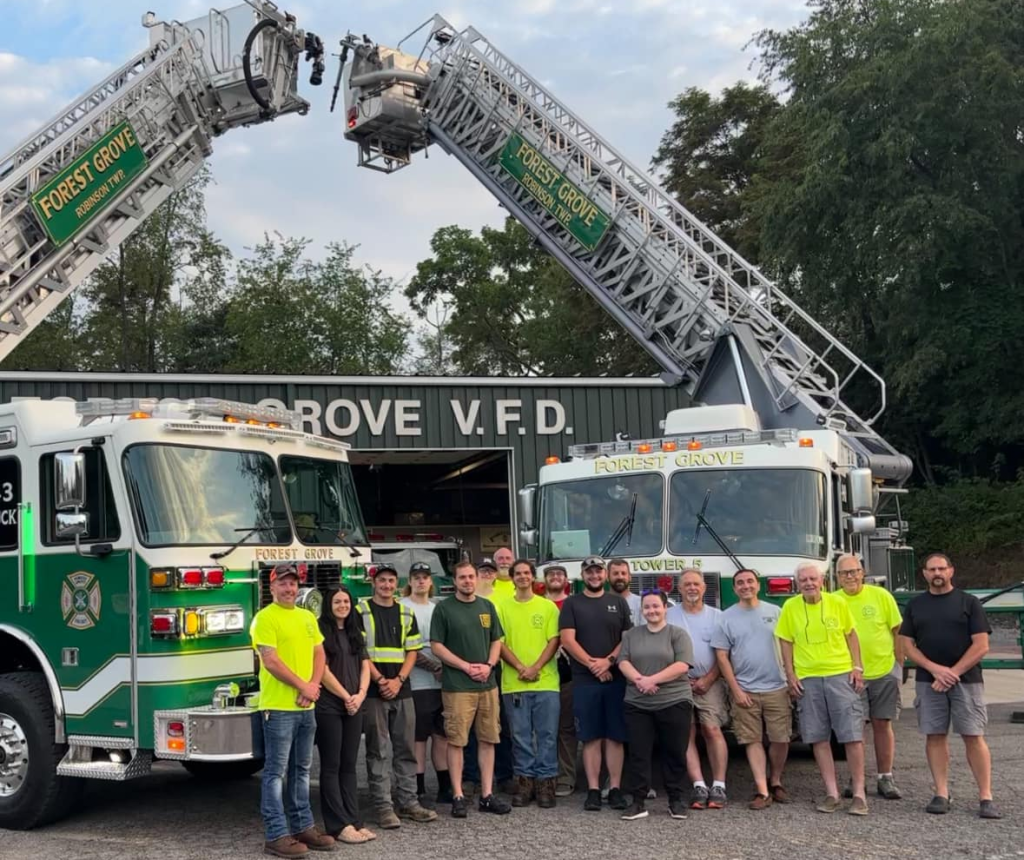 A group of people pose in front of two fire trucks and the Forest Grove V.F.D. fire station, with fire truck ladders raised in the background.