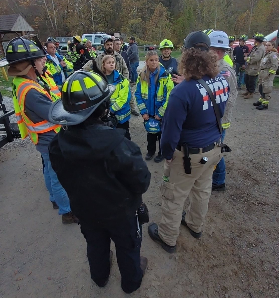 A group of emergency responders, wearing reflective vests and helmets, gather outdoors in a circle, listening to instructions. Trees and more responders are visible in the background.