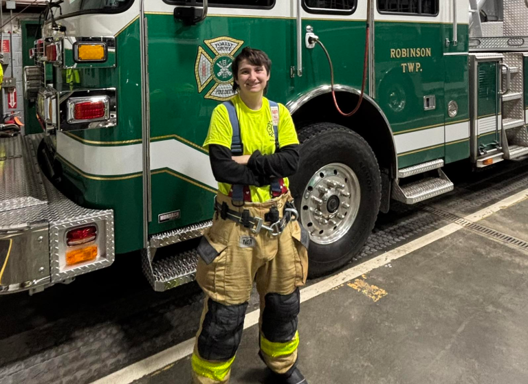 A Forest Grove new firefighter member in yellow gear and suspenders stands smiling with arms crossed in front of a green and white fire truck labeled “Robinson Twp.” inside a fire station.