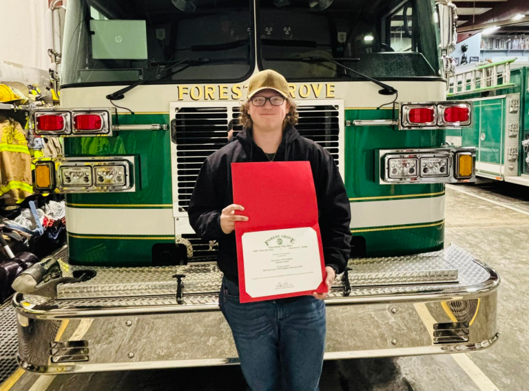 Person wearing glasses and a cap stands in front of a green fire truck, holding a red folder with a certificate inside. The truck’s front reads “Forest Grove,” and fire equipment is visible in the background, highlighting the proud moment as the Forest Grove firefighter of the year receives their recognition.
