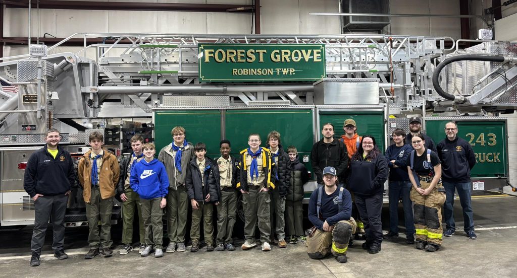 A group of scouts and firefighters pose in front of a large green fire truck labeled “Forest Grove Robinson TWP” inside a fire station.