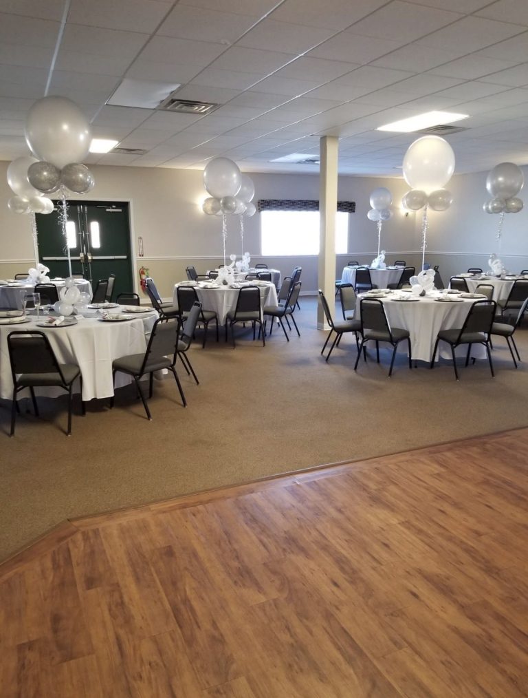 A banquet hall with round tables covered in white cloths, each decorated with silver and white balloon centerpieces. The chairs are arranged neatly around the tables on a tan carpeted floor, making this Forest Grove fire hall rental in Robinson Township an ideal setting for your next special event.