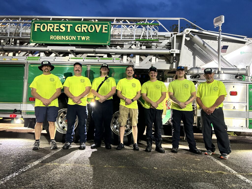 Seven firefighters in lime-green uniform shirts stand in a row to support the Forest Grove fire department in front of a ladder truck.